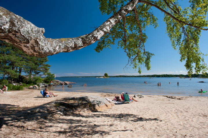 Utsikt över sandstranden där personer solar vid Vänerns vatten.