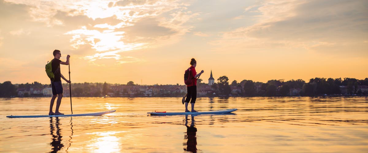 Två personer paddlar stand up paddling över en stilla Vättern i kvällssolen. 