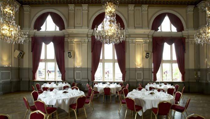 Banquet hall in an old building with crystal chandeliers in the ceiling, red velvet curtains in the large windows and white tablecloths at the round tables.