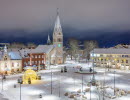 Vy över ett snötäckt Hertig Johans torg med kyrkan i bakgrunden och stämningsfull julbelysning. 