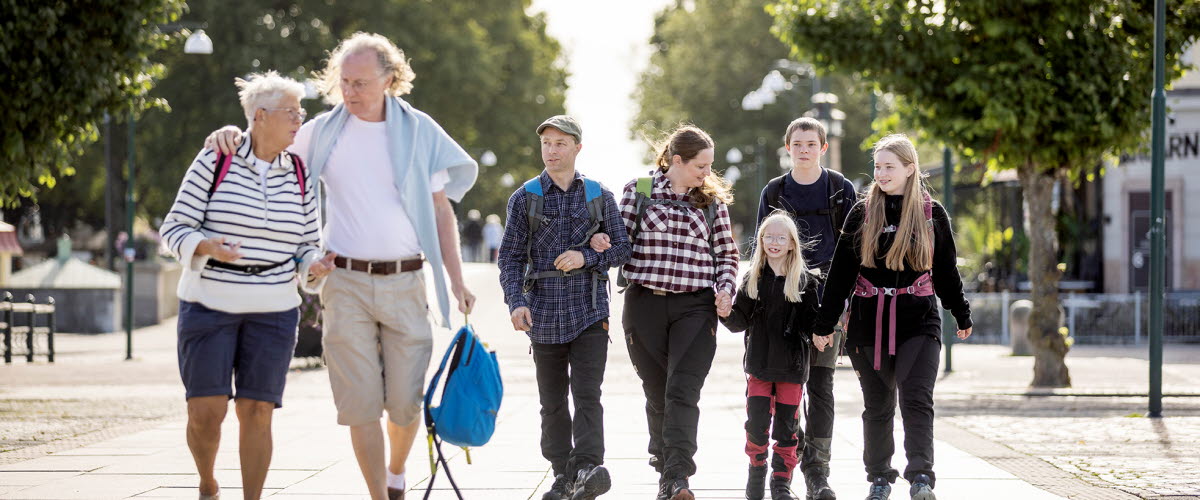 Ett äldre par och en familj med mamma, pappa och barn går på ett torg. Det finns gröna träd i bakgrunden.