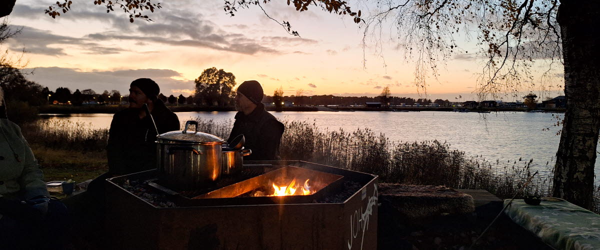 Två personer sitter på en bänk vid en tänd grillplats vid vatten. 