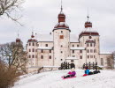 Två barn åker pulka från en backa framför slottet Läckö Slott. Marken är täck av snö. 