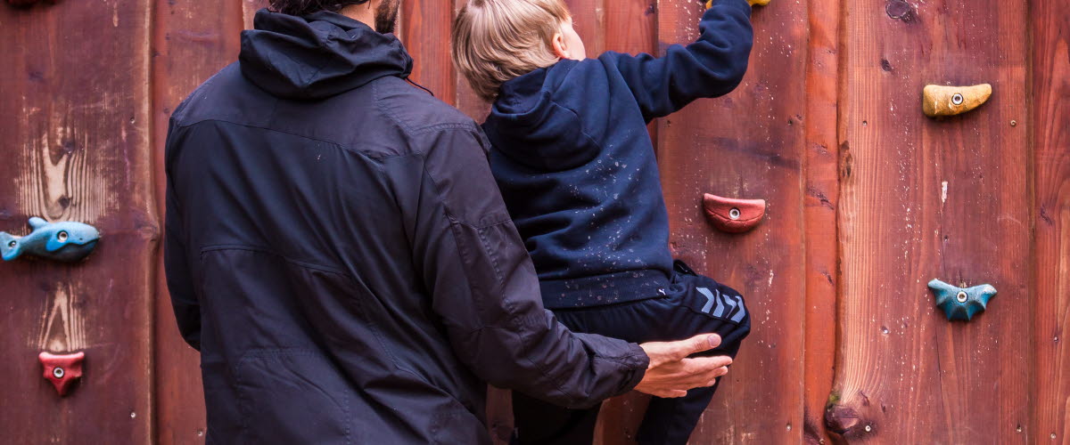 Ett litet barn klättrar på en klättervägg av trä. Barnets pappa stöttar barnet.