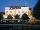 A large old white building with black roof right next to a river. It is evening and the lighting illuminates the building in a beautiful way.