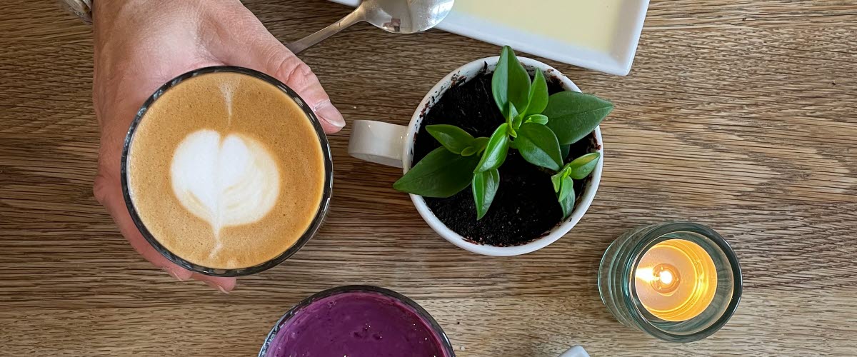 Ett bord med fat som det ligger kakor på. På bordet står också en kaffe latte och en smothie. Man kan se en hand som håller i glaset med kaffe latte och en hand som håller i en sked på väg att ta en bit av en kaka.