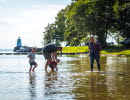 A family is wading in the water. The sun is shining and in the background you can se some trees an an old light house.