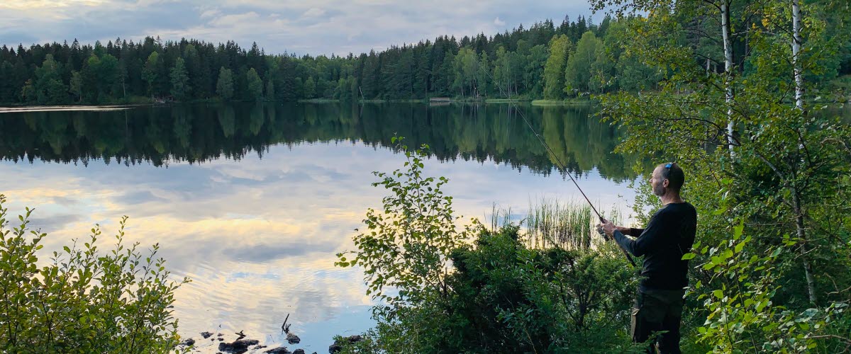 En man står vid kanten av en spegelblank sjö och fiskar i skymningen. 