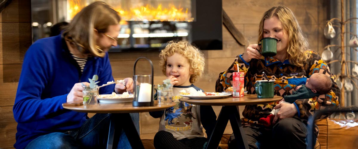 Pappa, son och mamma med liten bebis i famnen sitter framför brasan och äter och skrattar. 