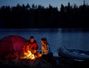 A man and a woman sitting by a bonfire outside their tent at night. Behind them there is a lake and a forest. Beside them there are a canoe.