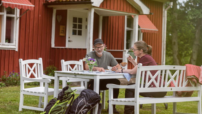 Röd stuga. Framför stugan sitter ett par och fikar i vita trädgårdsmöbler.