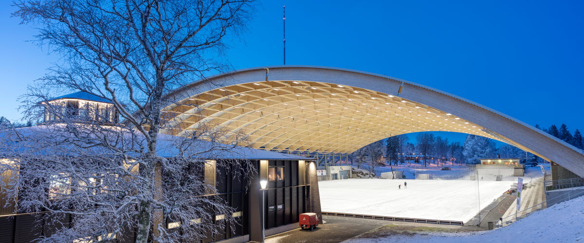 Upplyst svart byggnad(Billingecenter) i skymningen. Det liger snö på backen och i bakgrunden ser man det välvda taket över isbanan. 